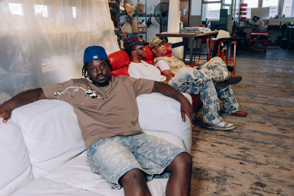 Group lifestyle shot in creative studio setting, featuring AXIS Studios Bamboo Panda Tee with panda embroidery on chest, styled with light wash patterned denim shorts, casual relaxed pose on white sofa.