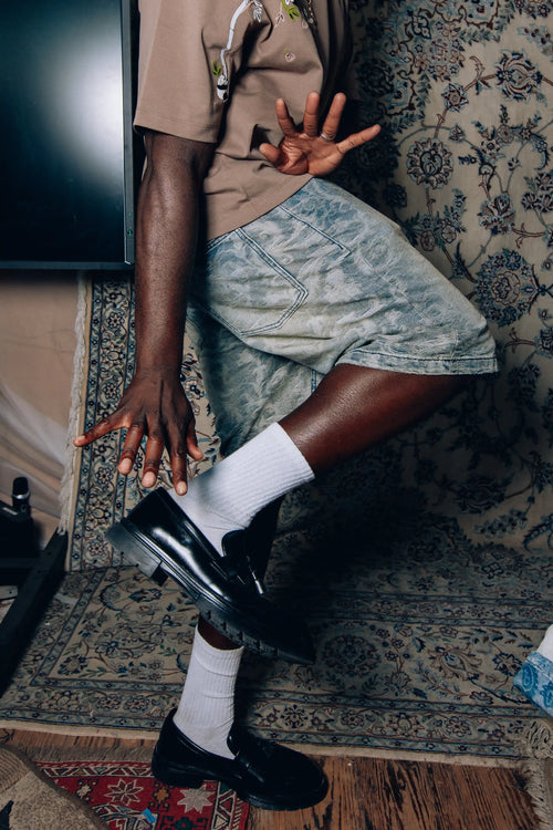 Model wearing Axis Studios washed cloud jacquard jorts with Panda Branch Tee, white socks and black loafers, ornate rug backdrop.