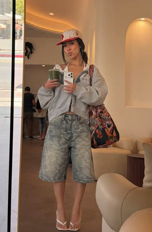 Woman wearing Axis Studios Cloud Jorts with cloud jacquard pattern, styled with grey zip-up hoodie and cap, mirror selfie in a café.